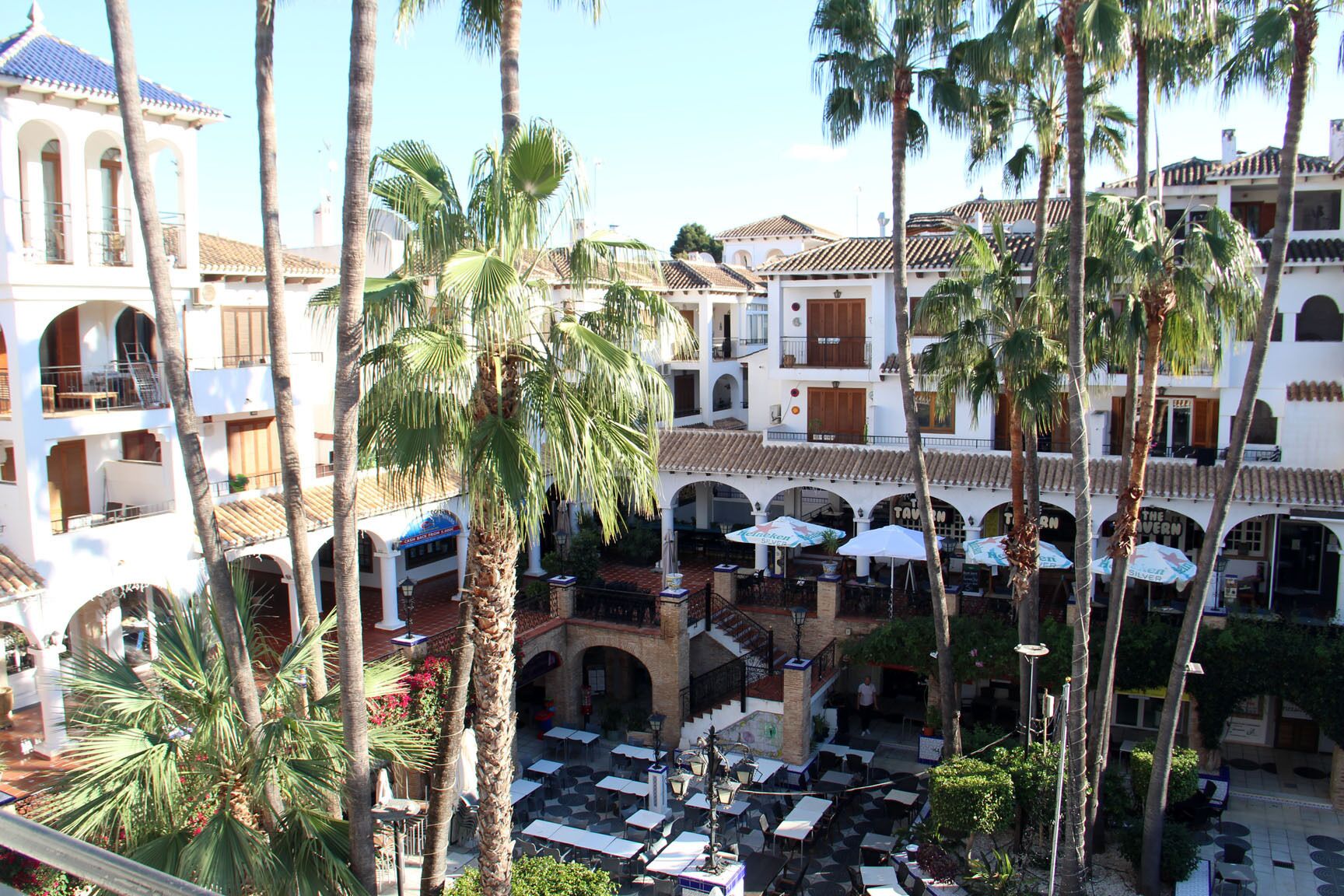 A courtyard with outdoor seating, palm trees, and buildings with tiled roofs.
