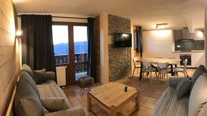 A cozy living room with a sofa, a coffee table, and a TV. There's a balcony with a view of mountains.