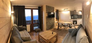 Una sala de estar acogedora con sofá, mesa de centro y televisor. Hay un balcón con vistas a montañas.