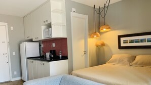 Een compacte hotelkamer met kitchenette, een bed met beige beddengoed, een grijze bank en een aan de muur bevestigd schilderij.