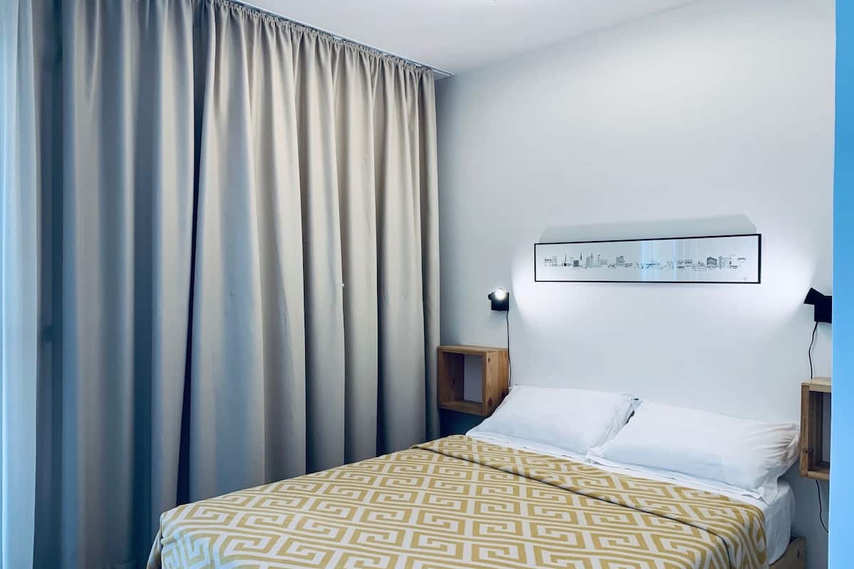 A hotel room with a bed, a headboard with a cityscape picture, a nightstand, and a window with curtains.