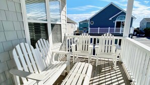 A covered porch with white Adirondack chairs and a small table, overlooking a blue house with a white fence. - Just steps to the beach! 🏝 Surf City Beach House (Surf City)