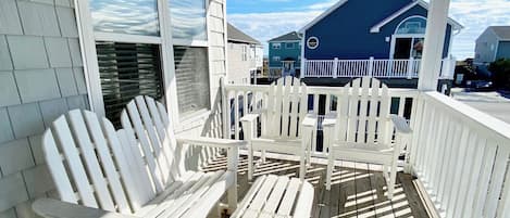 A covered porch with white Adirondack chairs and a small table, overlooking a blue house with a white fence.