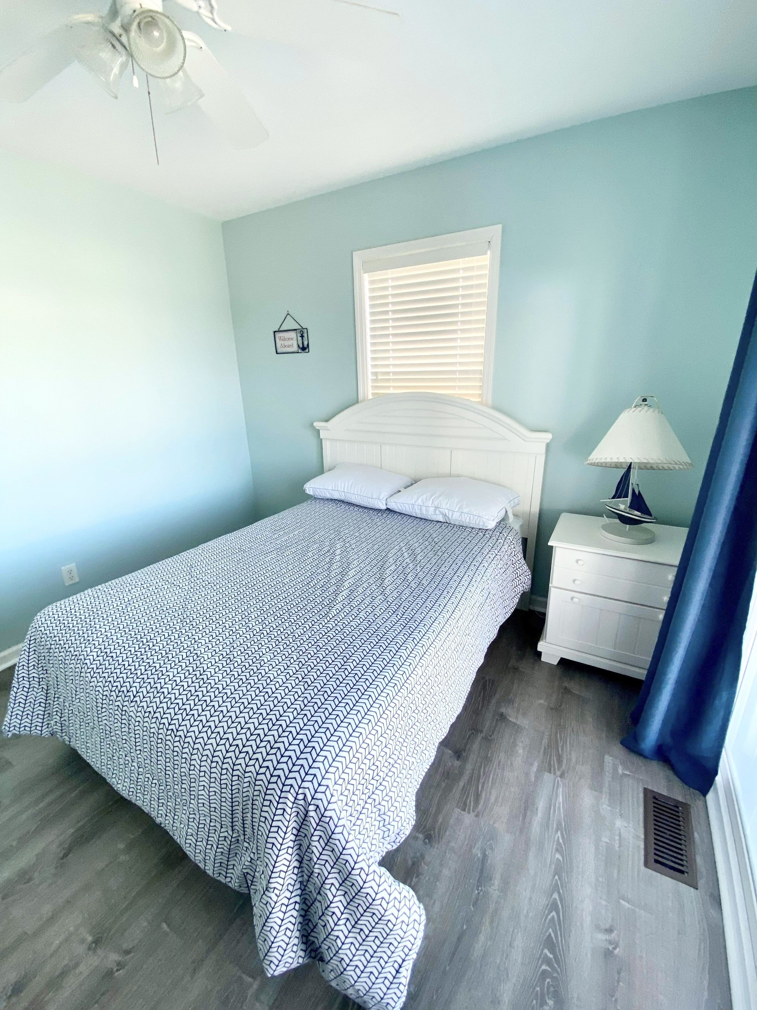 A bedroom with a bed, white nightstand, lamp, ceiling fan, window with blinds, and a wall-mounted light switch.