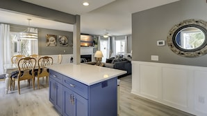 A modern kitchen with a blue island, dining area with wooden chairs, and a living room with a gray sofa.