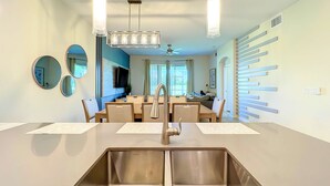 A modern kitchen with a stainless steel sink, a dining area with a table and chairs, and a living area with a couch and TV.