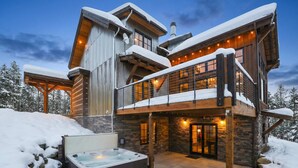 A two-story log cabin with a hot tub and stone walls, surrounded by snow.