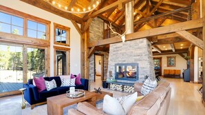 A spacious living room with a stone fireplace, wooden beams, and large windows overlooking a forested area.