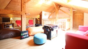 Een gezellige zolderkamer met een houten plafond, een rode stoel, een blauwe voetenbank, een spelletjes tafel en een televisie.