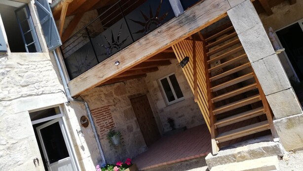 A two-story building with a wooden balcony, stone walls, and a small entrance area with a potted plant.