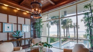 A modern living room with a large window overlooking a pool and palm trees, wooden paneling, and a hanging light fixture. - Aqua 3br/3ba. 2 Master BD ocean view (Panama City Beach)
