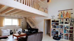 A spacious living room with a high ceiling, a staircase, a fireplace, and a large bookshelf.