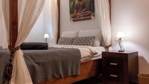 A bedroom with a bed, a nightstand, a lamp, a picture of a Buddha, and a curtain.