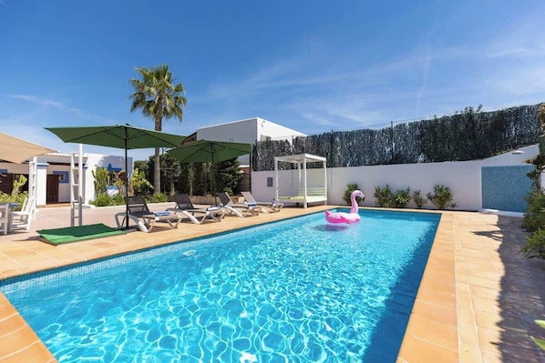 En swimmingpool med liggestole og parasoller, omgivet af en anlagt have og en moderne bygning.