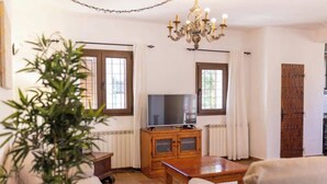 A living room with a television on a wooden cabinet, a chandelier, and a large window with curtains.