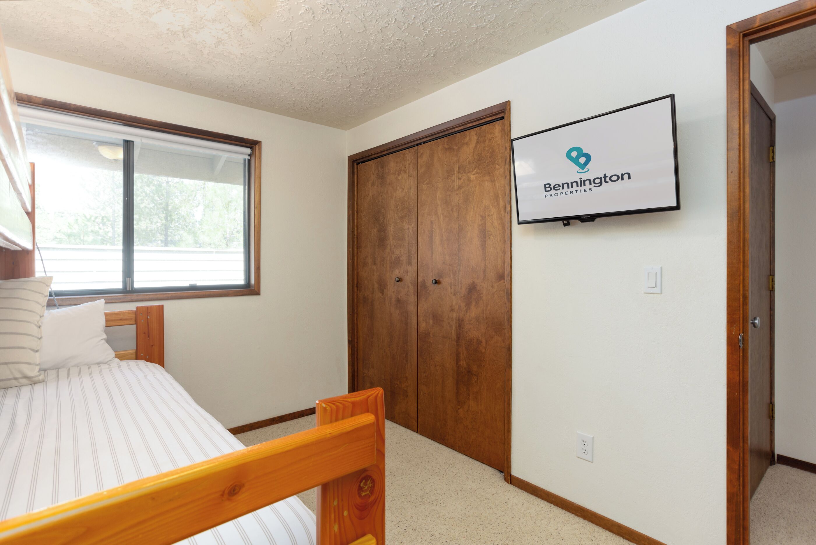 A bedroom with a bed, a wooden wardrobe, a window, and a wall-mounted television displaying 'Bennington Properties'.
