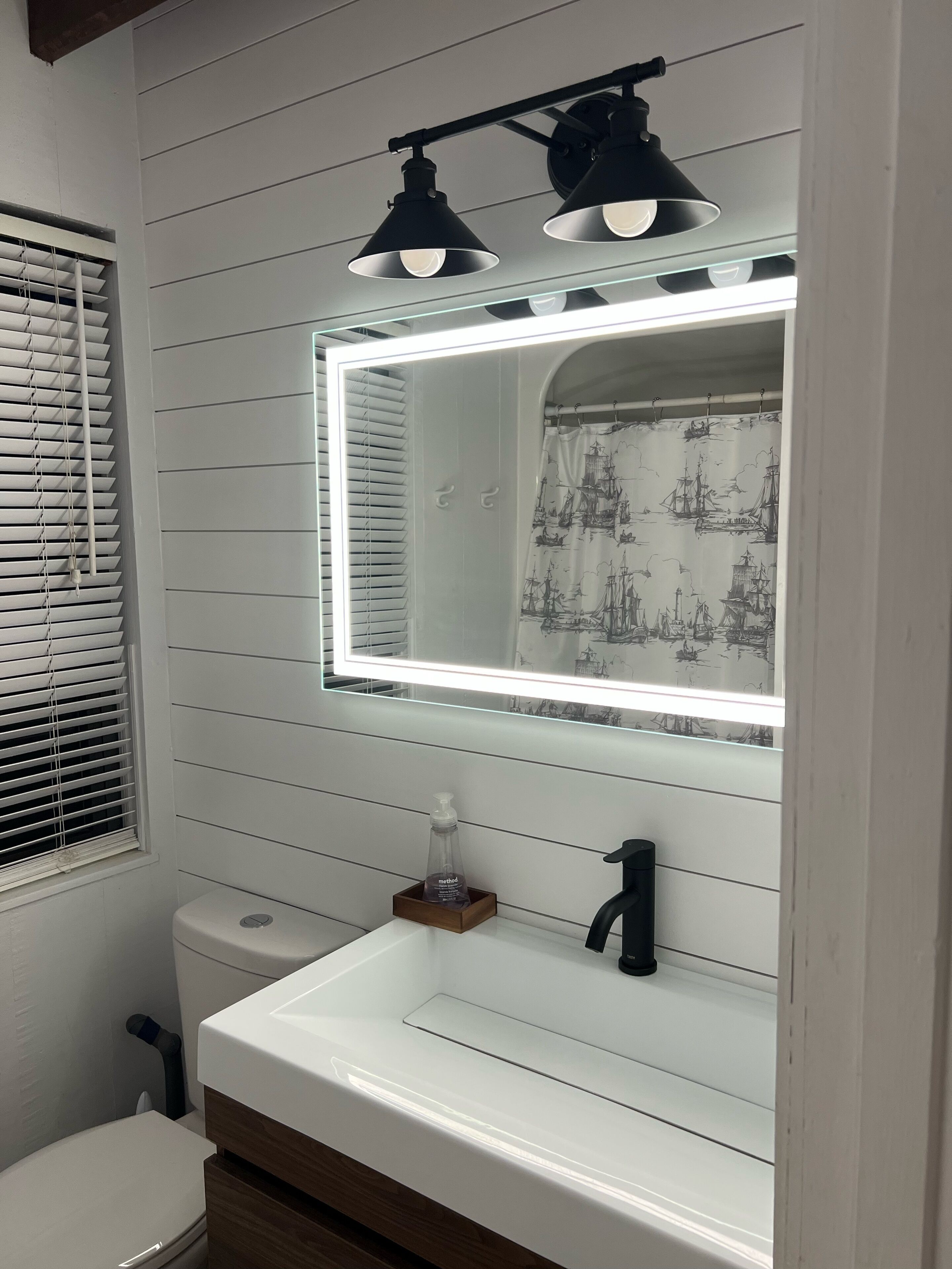 A bathroom with a white sink, a mirror with lights, and a window with blinds.