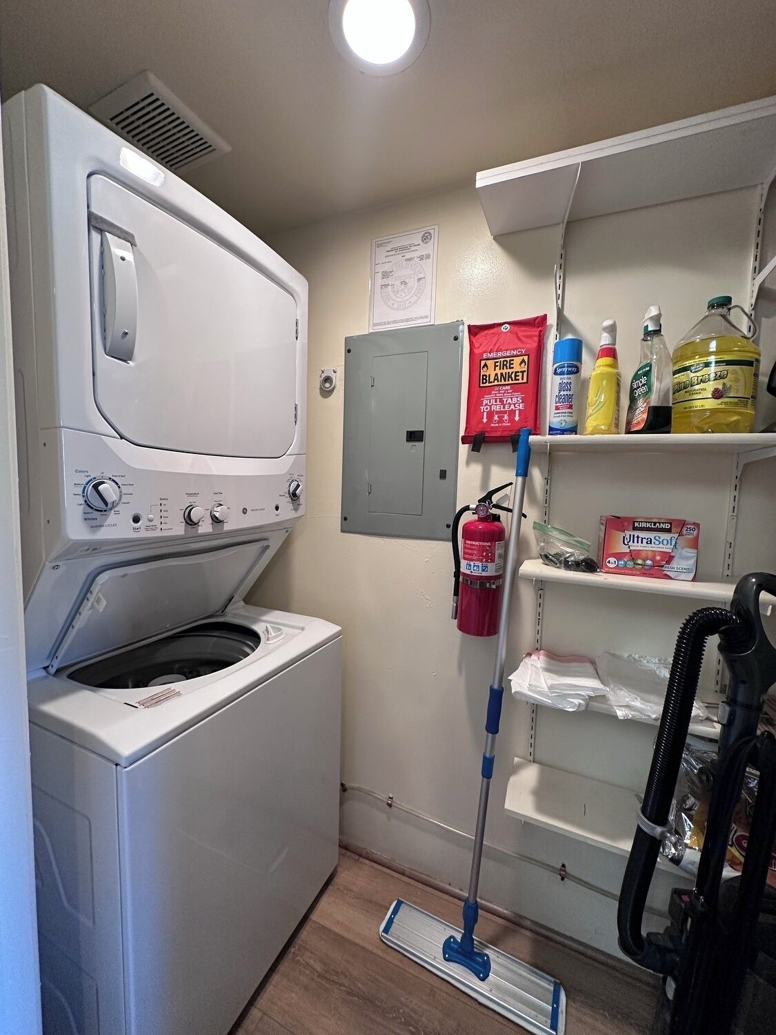 A laundry room with a front-loading washer and dryer, a fire extinguisher, cleaning supplies, and a vacuum cleaner.