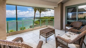 A balcony with wicker furniture, a small table, and a view of the ocean and palm trees.