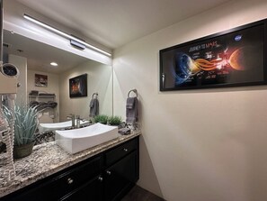 A modern bathroom with a granite countertop, a white sink, and a large mirror. There is a framed picture of a space mission on the wall.