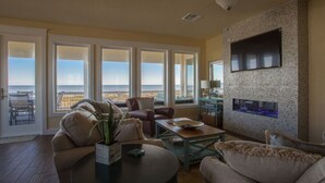 Ein geräumiges Wohnzimmer mit großem Fenster zum Meer, einem Kamin und einem an der Wand befestigten Fernseher.