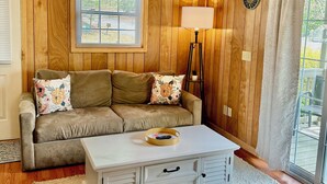 A cozy living room with wooden paneling, a sofa with floral cushions, a white coffee table, and a floor lamp. - Walking Distance to Lake James: Private Resort Amenities  (Marion)