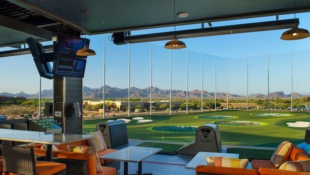 Blick auf einen Golfplatz von einer Innenlounge mit Korbmöbeln und einem TopGolf-Schild.