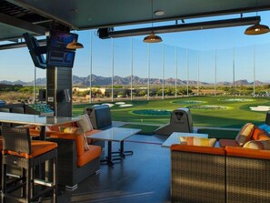 A golf course view from an indoor lounge with wicker furniture and a TopGolf sign. - ScottsdaleStays 8BDRM Million+ Home-Pool Spa-Theater-Golf & Bachelor HQ. (Scottsdale)