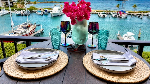 Mesa posta com pratos, talheres e copos, com vista para uma marina com barcos e céu claro.