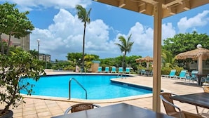 Piscina com espreguiçadeiras e guarda-sóis, rodeada por vegetação e edifícios.