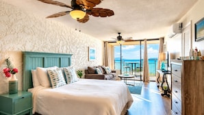 Quarto com cama, mesa de cabeceira, cômoda, ventilador de teto e vista para o oceano.