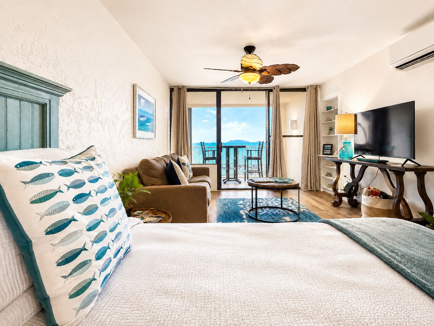 Una camera d'albergo con un letto, un divano, una TV, una ventola a soffitto e un balcone con vista.