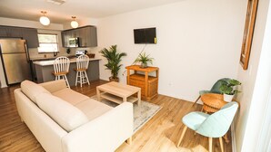 A modern living room with a sofa, a coffee table, and a dining area with chairs. There is a kitchen in the background with a refrigerator and a TV mounted on the wall. - NEWLY RENOVATED SOPHISTICATED INDUSTRIAL TOWN HOME (St. Petersburg)