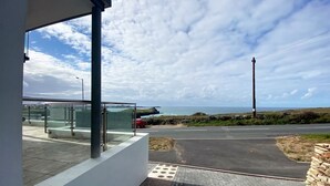 Un bâtiment moderne doté d’un balcon en verre donnant sur une zone pavée et une route menant à la mer.