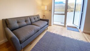 Un canapé gris dans une pièce dotée d’un plancher en bois, d’un tapis à motifs, d’une lampe et d’une porte-fenêtre coulissante donnant sur un espace extérieur délimité par une clôture en bois.