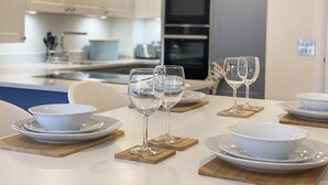 Une cuisine moderne avec des armoires blanches, un four encastré et un coin repas agrémenté de bols blancs et de verres à vin posés sur des sous-verres en bois.