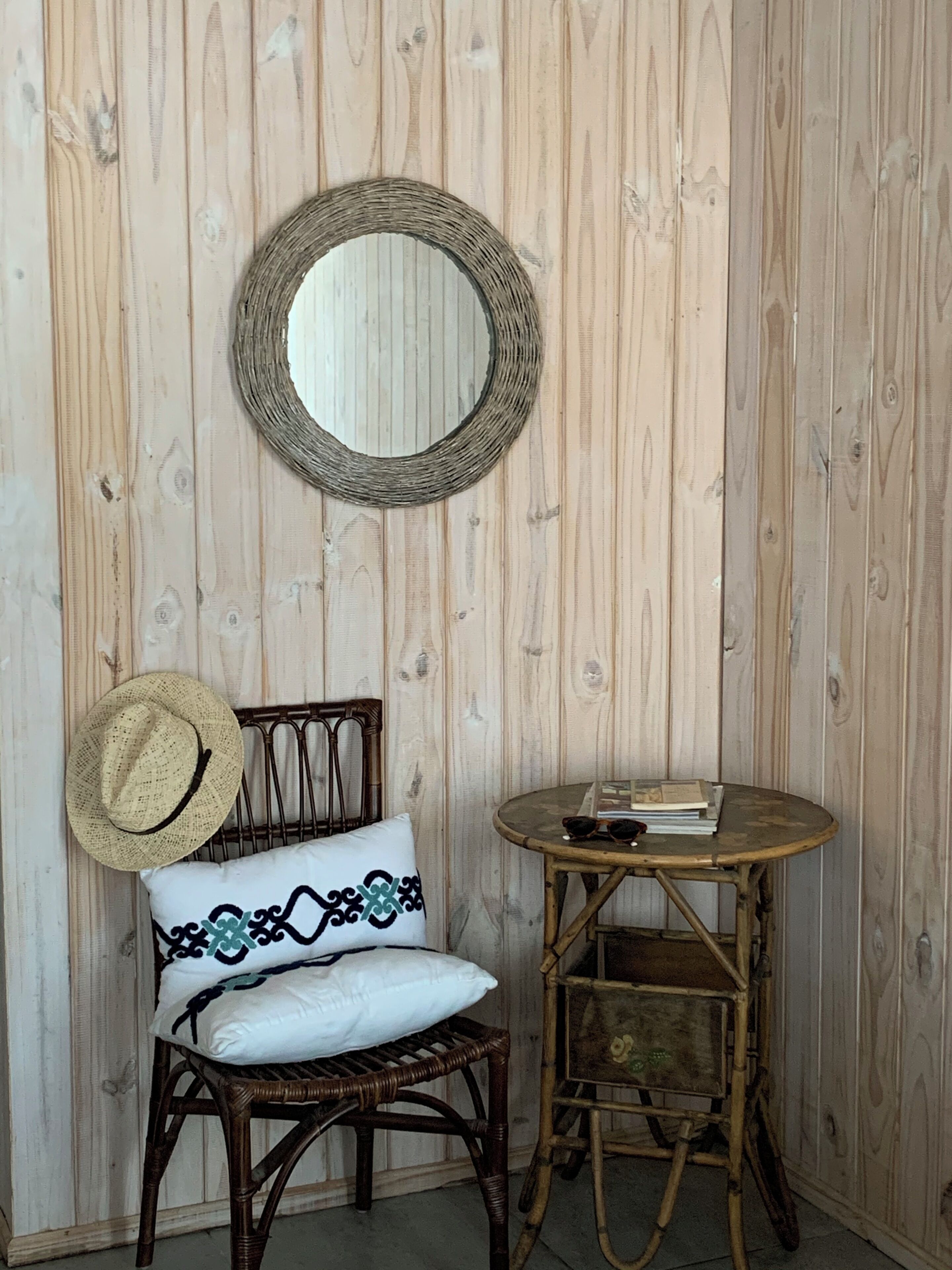 Un rincón con una silla de mimbre, un espejo redondo, una mesita redonda con libros y gafas de sol, y un cojín decorativo.