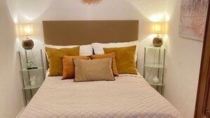 Un lit bien fait, recouvert d’une courtepointe et agrémenté de plusieurs oreillers, flanqué de deux tables de chevet avec des lampes, d’un objet décoratif fixé au mur et d’une armoire en bois.