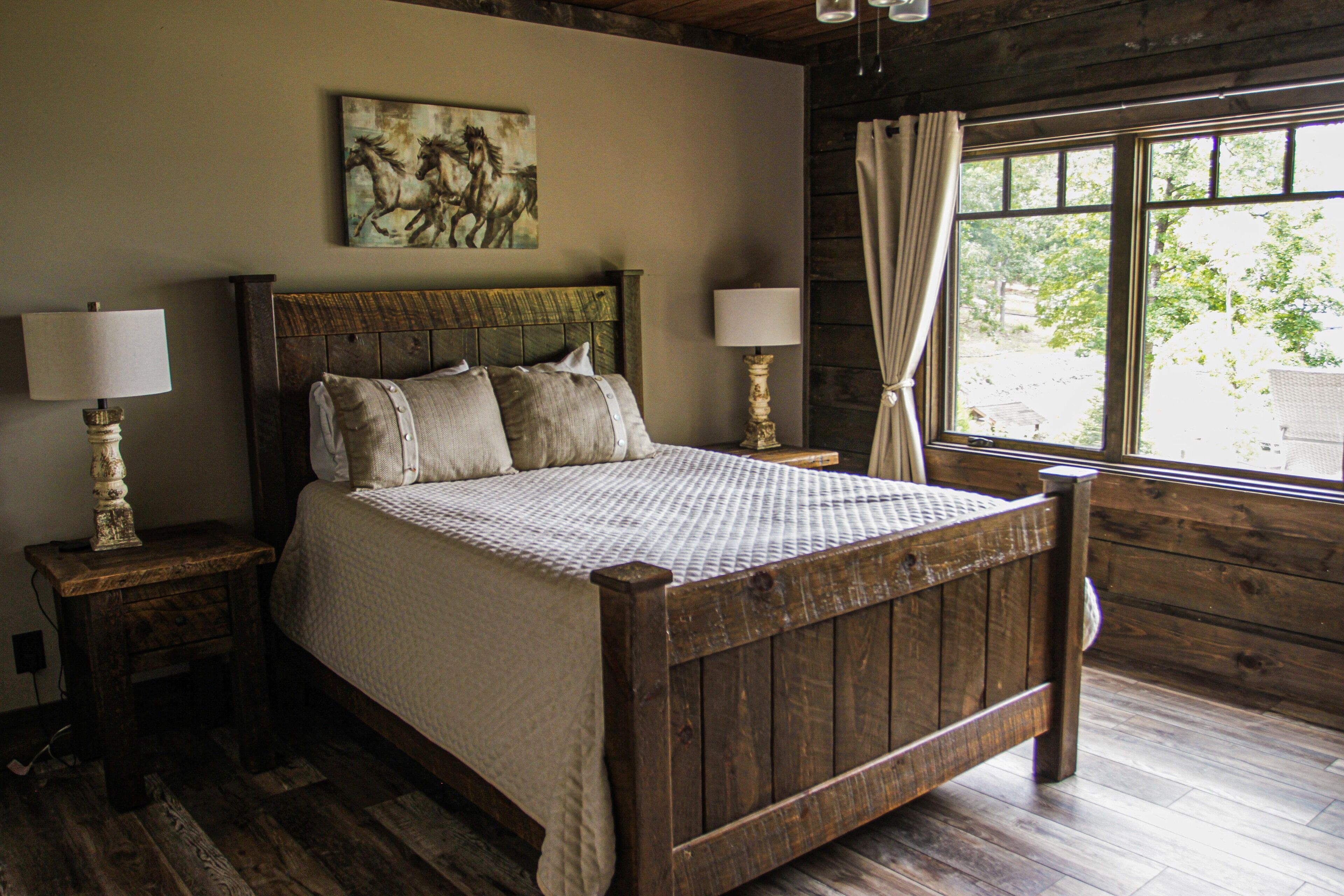 A wooden four-poster bed with a quilted bedspread, two bedside tables with lamps, a window with curtains, and a painting of horses on the wall.