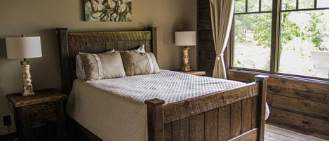 A wooden four-poster bed with a quilted bedspread, two bedside tables with lamps, a window with curtains, and a painting of horses on the wall.