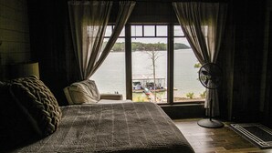 Een slaapkamer met een groot raam dat uitkijkt op een wateroppervlak, een bed, een stoel en een ventilator.