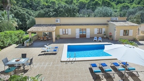Zona de piscina con sillones de descanso y una mesa, rodeada por un edificio con tejado de azulejos y vistas a una ladera boscosa.