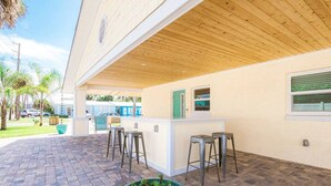 Une terrasse extérieure dotée d’un plafond en bois, d’un comptoir blanc avec des tabourets et d’une grande plante en pot.