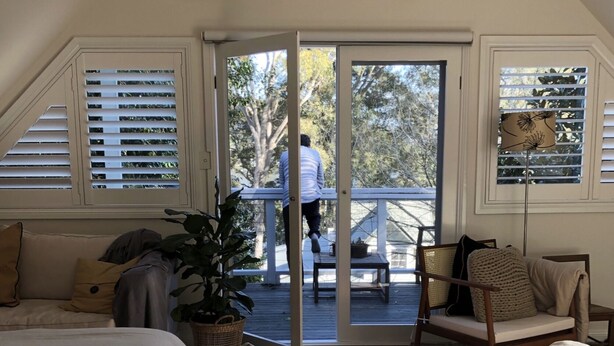 Une chambre avec un grand lit, une plante en pot, un plancher en bois et une porte-fenêtre coulissante donnant sur un balcon.
