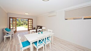 A dining area with a white table and chairs, a sofa, and a view of a garden through glass doors. - Close to beach and town in a quiet location.  (Busselton)