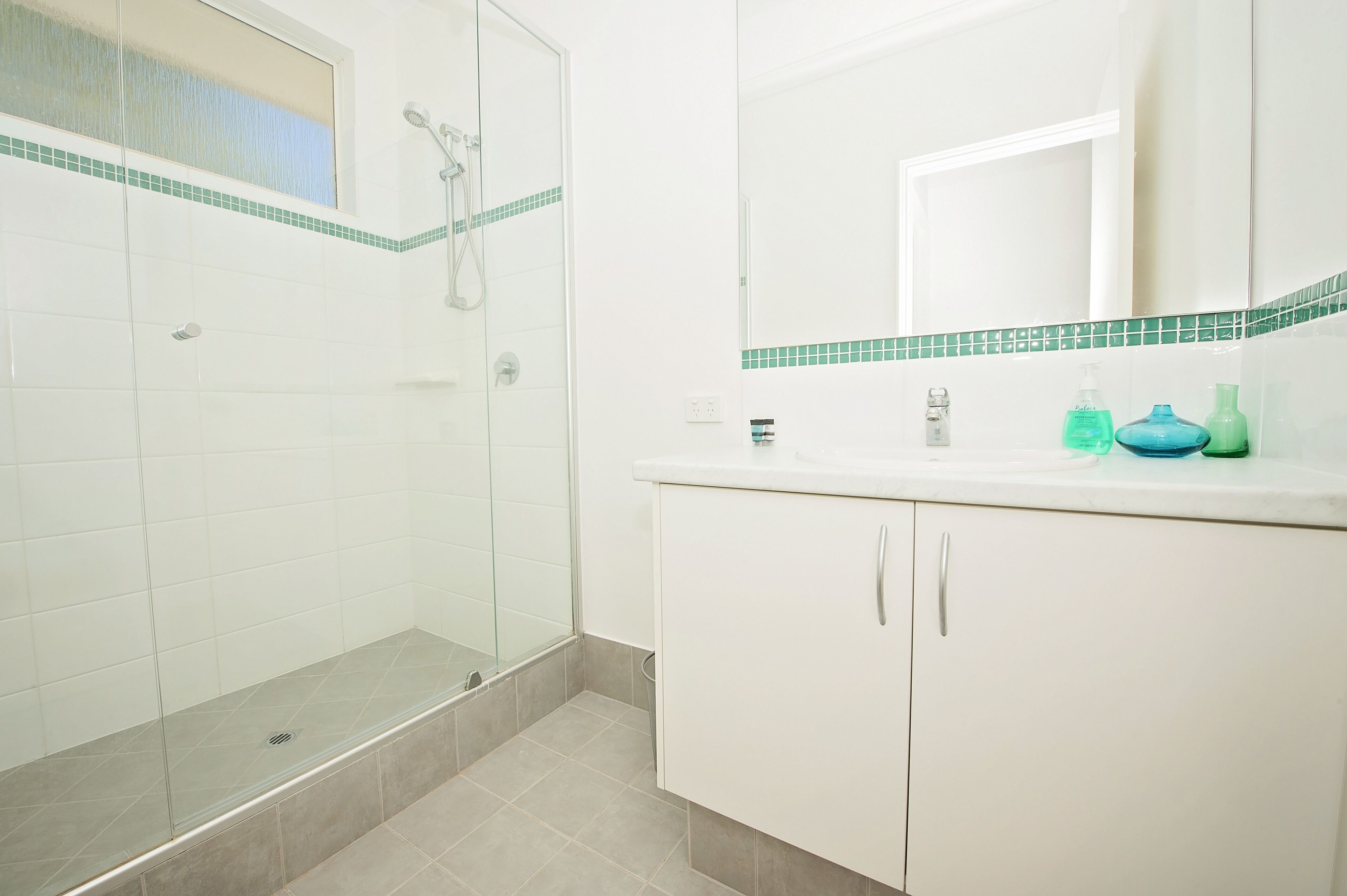 A bathroom with a glass-enclosed shower, white tiled walls, a mirror, and a white vanity with a green-tiled countertop.
