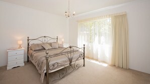 A bedroom with a metal bed frame, a bedside table with a lamp, and a window with sheer curtains. - Close to beach and town in a quiet location.  (Busselton)