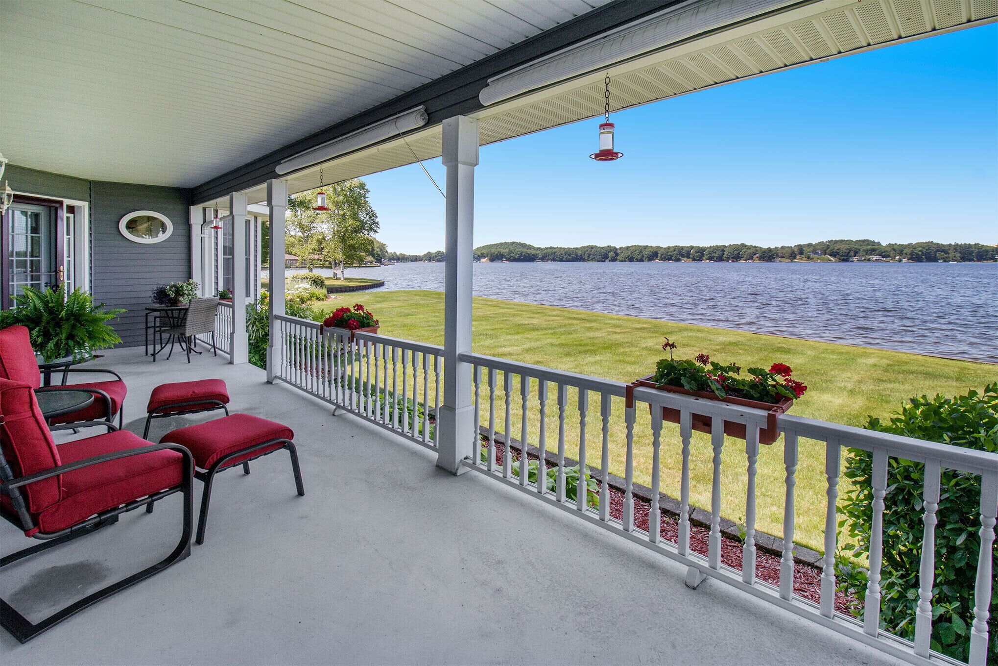 A covered porch with red chairs and a table, overlooking a lake with a grassy area and a white fence.