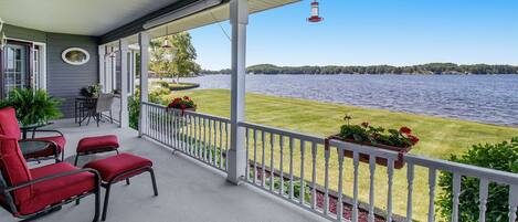 A covered porch with red chairs and a table, overlooking a lake with a grassy area and a white fence.