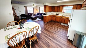 A spacious living area with a dining table set for four, wooden flooring, a kitchen with wooden cabinets, and a ceiling fan.
